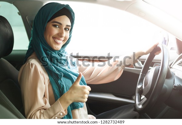 Happy arabian woman wearing\
hijab driving her car with thumbs up gesture. Get driving license\
concept