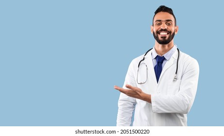 Happy Arabic Doctor Man Smiling Showing Copy Space With Hand Advertising Your Text Over Blue Studio Background. Portrait Of Successful Physician. Medical Advertisement Banner