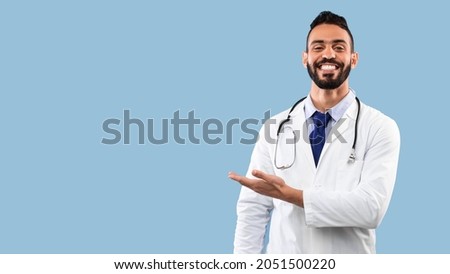 Happy Arabic Doctor Man Smiling Showing Copy Space With Hand Advertising Your Text Over Blue Studio Background. Portrait Of Successful Physician. Medical Advertisement Banner