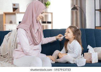 Happy Arabic Family, Morning Hygiene. Muslim Mother In Hijab, Sitting On Sofa At Home, Brushing Teeth Of Her Cute Little Daughter Child With Electric Toothbrush, Showing How To Clean Teeth Propelry.