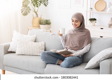Happy Arabic Housewife In Hijab Relaxing With Book And Coffee On Couch At Home, Enjoying Indoor Leisure, Resting On Comfortable Sofa, Free Space