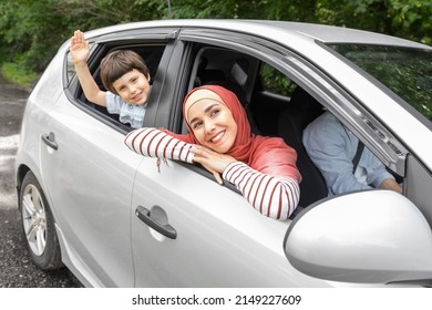 Happy Arabic Wife In Hijab And Child Waving Hands In Open Window At Car, Husband At Steering Wheel, Outdoors In Summer. Family Travel, People Enjoy Weekend, Vacation At New Auto And Life Insurance