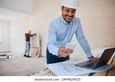 Happy architect texting on mobile phone while using laptop and analyzing blueprints at construction site. Copy space.  - Powered by Shutterstock