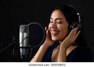 Happy Asain Woman As Beautiful Professional Singer Catching Earphone, Smile, Enjoy Practicing Attractive Voice And Song Singing Performance To Microphone In Dark Sound Record Room Of Music Studio