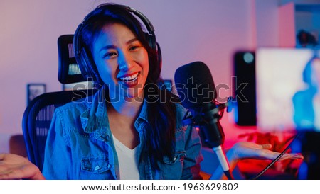 Happy asia girl blogger music influencer use smartphone broadcast recording wear headphone online live talk with listening audience in living room home studio at night. Content creator concept.