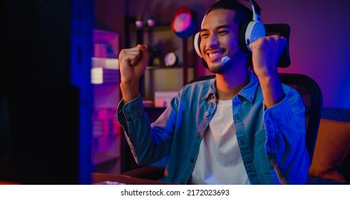 Happy asia guy gamer wear headphone participation play video game colorful neon lights computer in living room at night modern house. Esport streaming game online, Home quarantine activity concept. - Shutterstock ID 2172023693