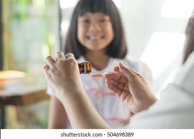 Happy Asia Little Child Girl Smiling Want To Take Medicine Form Mother. Asian Kid Female Waiting To Eat Drug. Hands Of Mom Pouring Cough Syrup Medicine Into Clear Spoon To Daughter.