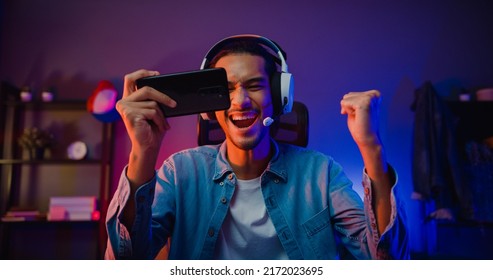 Happy asia man gamer wear headphone competition play video game online with smartphone colorful neon light in living room at night modern house. Esport streaming game online, Home quarantine activity. - Shutterstock ID 2172023695