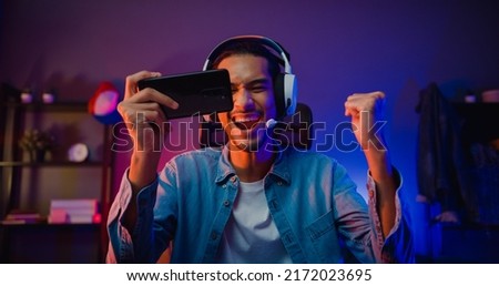 Happy asia man gamer wear headphone competition play video game online with smartphone colorful neon light in living room at night modern house. Esport streaming game online, Home quarantine activity. Stock foto © 
