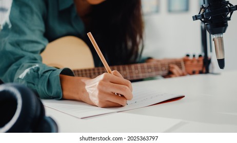 Happy asia woman songwriter play acoustic guitar listen song from smartphone think and write notes lyrics song in paper sit in living room at home studio. Music production at home concept.