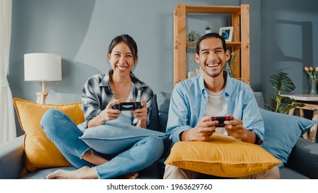 Happy Asia Young Couple Man And Woman Sit On Couch Use Joystick Controller Play Video Game Spend Fun Time Together In Living Room. Asian Married Couple Family Lifestyle, Couple Stay At Home Concept.