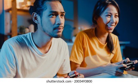 Happy Asia Young Couple Man And Woman Sit Couch Use Joystick Controller Play Video Game Spend Fun Time Together In Living Room At Night. Asian Married Couple Family Lifestyle, Stay At Home Concept.