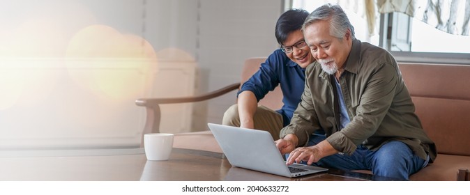 Happy Asian Adult Son And Senior Father Sitting On Sofa Using Laptop Together At Home . Young Man Teaching Old Dad Using Internet Online With Computer On Couch In Living Room . Copy Space Banner
