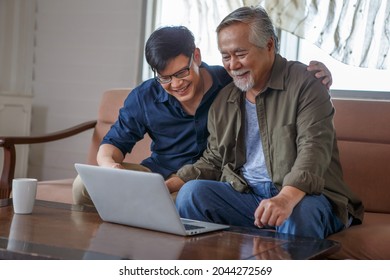 Happy Asian Adult Son And Senior Father Sitting On Sofa Using Laptop Together At Home . Young Man Teaching Old Dad Using Internet Online With Computer On Couch In Living Room . 