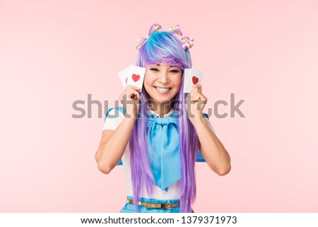 Happy asian anime girl in purple wig holding cards with hearts isolated on pink