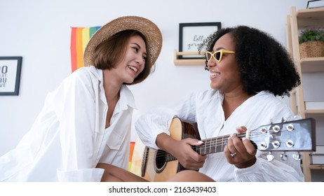 Happy asian Arican couple lesbian woman playing acoustic guitar sitting together on sofa at home. Lgbtq lover girl play song music with guitar enjoying activity hobby leisure in bed room. - Powered by Shutterstock