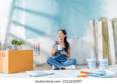 Happy Asian attractive young woman having coffee break during home renovation her living room. Woman repairman painted the wall with paint makes repairs and sits on a floor home relocation renovate - Powered by Shutterstock