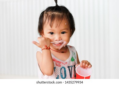 Happy Asian Baby Child Girl Aged Of 1 Year And 4 Months Olds Eating Yogurt Food By Herself. Her Face Is Sloppy And Messy. Kid And Food Concept. On White Background.