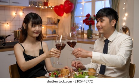 Happy Asian Beautiful Couple Lifting Wine Glasses And Toasting For Valentine’s Day While Having Romantic Dinner At Home With Light Decors