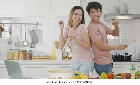 Happy Asian Beautiful Family Couple Husband And Wife Singing In Kitchenware Microphones In Kitchen Together Having Fun Dance Listen Music At Home. Two People Dancing