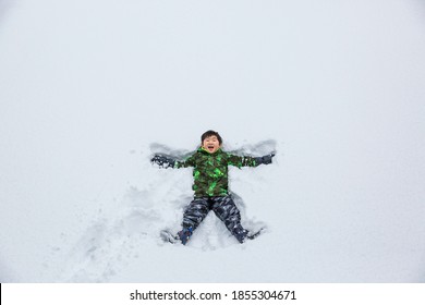 A Happy Asian Boy Making Snow Angel On A White Snow Field.