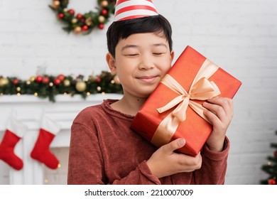 Happy Asian Boy In Party Cap Holding Gift Near Blurred Christmas Decor At Home