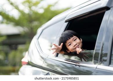 Happy Asian Boy Waving Hands Gesturing Hello Out Of The Car Window During A Trip With His Family. Little Child Sticking Head Outta The Windshield Traveling In A Car On A Summer Vacation.