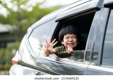 Happy Asian Boy Waving Hands Gesturing Hello Out Of The Car Window During A Trip With His Family. Little Child Sticking Head Outta The Windshield Traveling In A Car On A Summer Vacation.