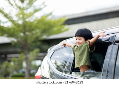 Happy Asian Boy Waving Hands Gesturing Hello Out Of The Car Window During A Trip With His Family. Little Child Sticking Head Outta The Windshield Traveling In A Car On A Summer Vacation.