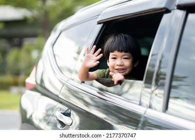 Happy Asian Boy Waving Hands Gesturing Hello Out Of The Car Window During A Trip With His Family. Little Child Sticking Head Outta The Windshield Traveling In A Car On A Summer Vacation.