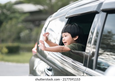 Happy Asian Boy Waving Hands Gesturing Hello Out Of The Car Window During A Trip With His Family. Little Child Sticking Head Outta The Windshield Traveling In A Car On A Summer Vacation.