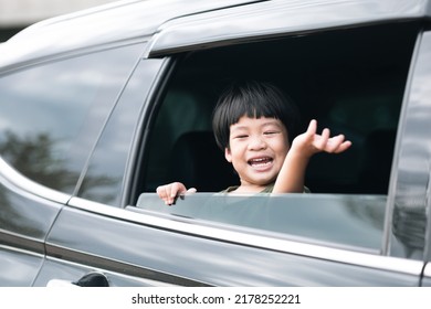 Happy Asian Boy Waving Hands Gesturing Hello Out Of The Car Window During A Trip With His Family. Little Child Sticking Head Outta The Windshield Traveling In A Car On A Summer Vacation.