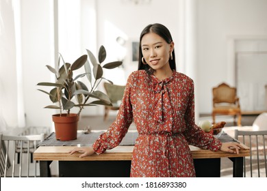 Happy Asian brunette woman in red stylish floral dress smiles, looks into camera and leans on wooden table at kitchen. - Powered by Shutterstock