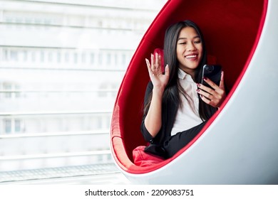 Happy Asian Business Woman Sitting Inside Ball Chair At Office Waving During Video Call