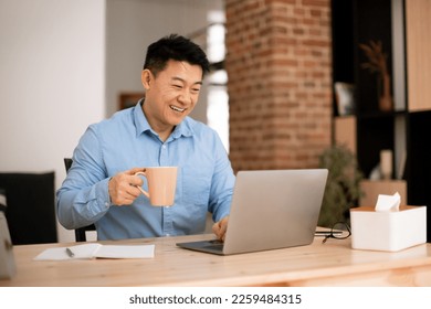 Happy asian businessman drinking coffee while using laptop, sitting during break with cup at workplace at home office. Successful entrepreneurship and freelance career - Powered by Shutterstock