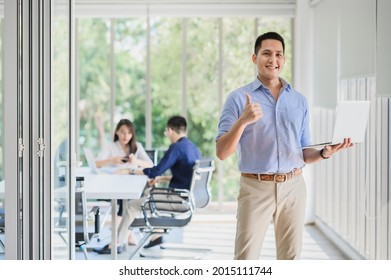 Happy Asian businessman holding laptop and smiling with thumbs up gesture with colleague in background. Business success concept
