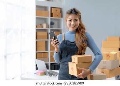 Happy Asian businesswoman starting sme holding smartphone. Small Online Business Owner, SME Work From Home Box, SME Online Marketing and Product Packing and Delivery Service. - Powered by Shutterstock