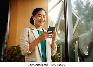 Happy Asian businesswoman text messaging on cell phone while standing by the window in the office.