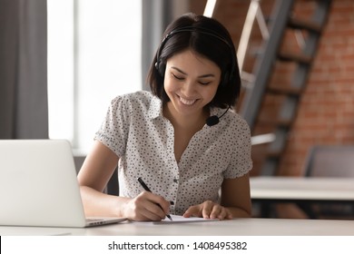 Happy asian businesswoman wearing headset make notes talk by conference call doing video chat on laptop in office, smiling chinese support service agent write information consult customer