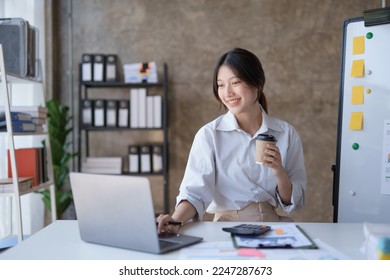 Happy Asian businesswoman working and checking work with laptop computer while having a comfortable cup of coffee at office.