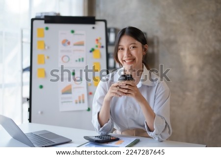 Happy Asian businesswoman working and checking work with laptop computer while having a comfortable cup of coffee at office.