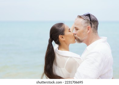 Happy Asian and caucasian white couple enjoy relaxing together at the beach during vacation or their honeymoon dating. Husband gently kissing his wife. - Shutterstock ID 2231706695