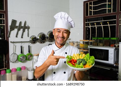 Happy Asian Chef Showing Plate With Raw Vegetables On The Kitchen. Selective Focus.