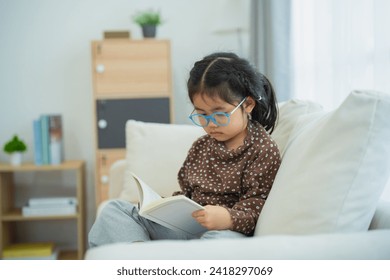 Happy asian child baby girl smiling wearing glasses and reading book while sitting on couch sofa in living room at home. Girl relex reading book smile at sofa In the house. Back to school concept. - Powered by Shutterstock