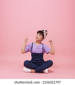 Happy Asian Child Girl Pointing Fingers Up On Pink Background With Copy Space. 