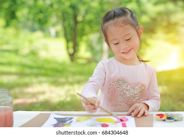 Happy Asian Child Girl Sitting At A Table In A Summer Garden Painting With Paintbrush, Education Art Concept.