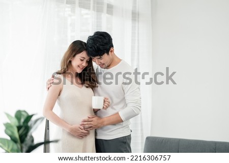 A happy asian couple. A beautiful pregnant wife is holding a cup of milk for the baby with her husband smiling and both of their hands rubbing the wife's tummy in the living room.