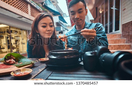 Happy asian couple enjoy chinese food. Couple is siting at cafe or restaurant. They have casual style - denim jackets. There are camera on the table.