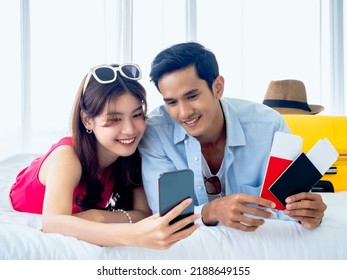 Happy Asian Couple With Flight Booking Or Shopping Online For Summer Trip, Travel Abroad. Young Man And Woman Looking At Smartphone And Holding Two Passports And Tickets While Lying In Bed Together.