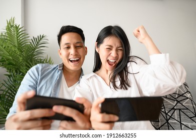 Happy Asian Couple Having Fun Playing Game In Smartphone Mobile In Living Room